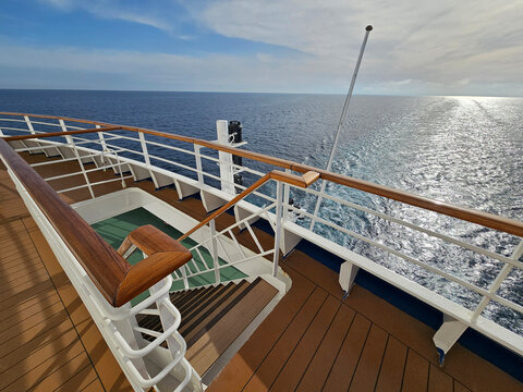 Sunlight glints on calm ocean waters beyond a cruise ship deck. Wooden railings and stairs frame the view from a ship upper level. Clear sky with gentle clouds above the sparkling sea surface