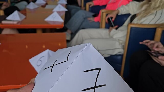 Hand holds paper pyramid with handwritten math problem or game Mafia. Other pyramids rest on orange tables. Audience seated in background, blurred. Simple educational activity, classroom setting