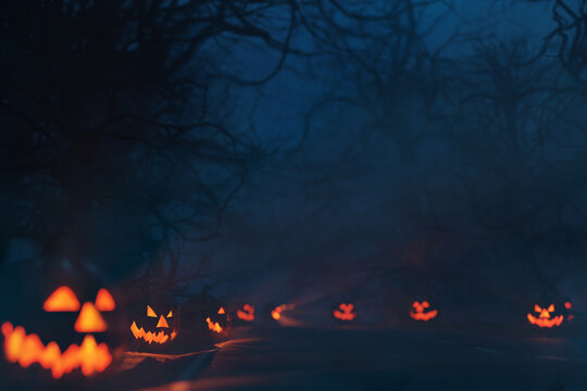 Misty spooky forest path at night, lined with glowing carved Jack-o'-lanterns. Bare, twisted trees and thick fog create a haunting Halloween scene. Scary road at night. Halloween background