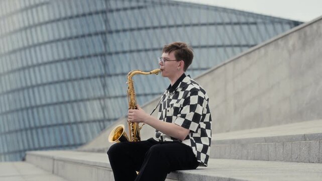 Caucasian young adult musician playing saxophone outdoors on city steps in slow motion. Footage ideal for urban culture marketing, music festivals and youth arts advertising