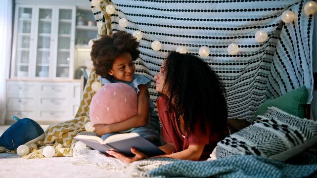 Girl hugging pillow listening mom reading book blanket fort. Cozy home evening.