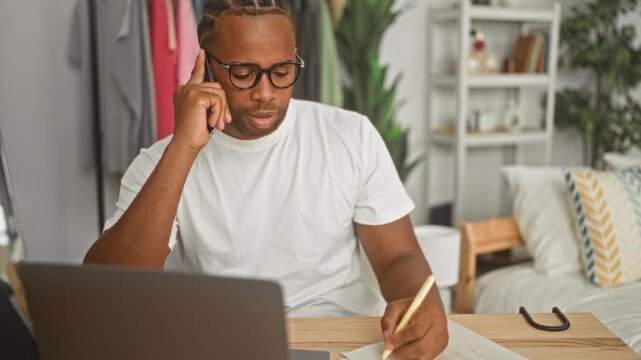 Man holding phone to ear while writing notes at a laptop in building; concentration remote work productivity.