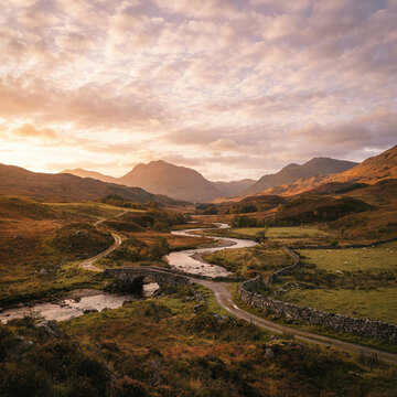 A stone bridge crosses a winding river through a mountain valley as sheep graze nearby