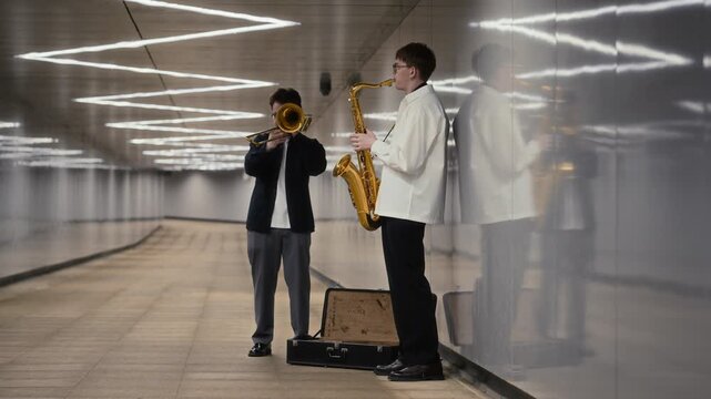 Two young adult Caucasian men playing saxophone and trombone in modern urban pedestrian tunnel with open case. Footage suitable for youth culture, creative arts and street performance projects