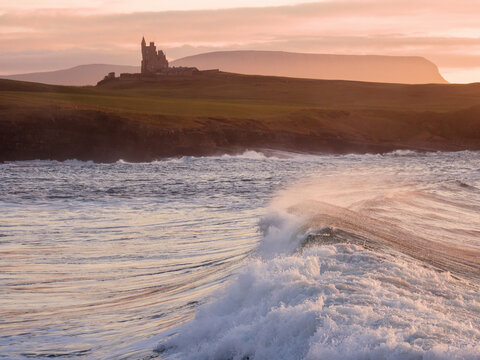 Dramatic coastal scene at sunset, waves crash near a cliff. Ancient stone castle perches on distant green hills. Warm golden light bathes the landscape. Castle Classiebawn Viewpoint, Ireland