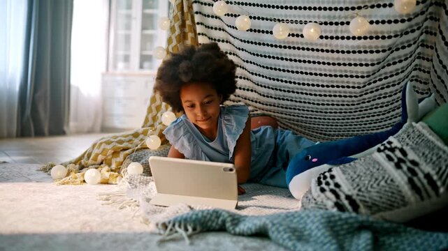 Kid using tablet lying blanket fort. African american girl smiling cozy home.