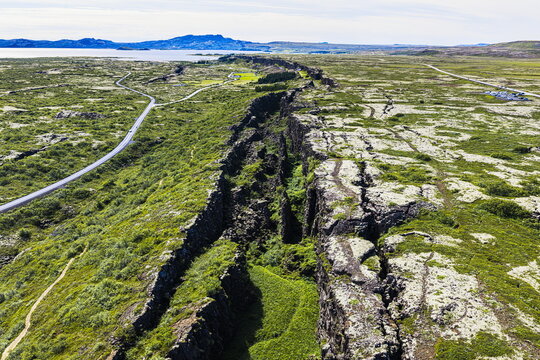 Aerial view of Almannagja gorge featuring dramatic tectonic rifts, moss-covered lava fields, and a winding road through the rugged landscape of Blaskogabyggd, Iceland.