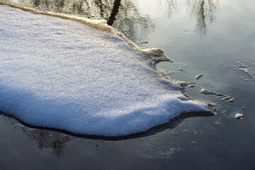 Close-up of sparkling snow texture on thick ice crust floating in dark cold river water during early spring thaw, soft natural lighting at sunset © PhotoChur