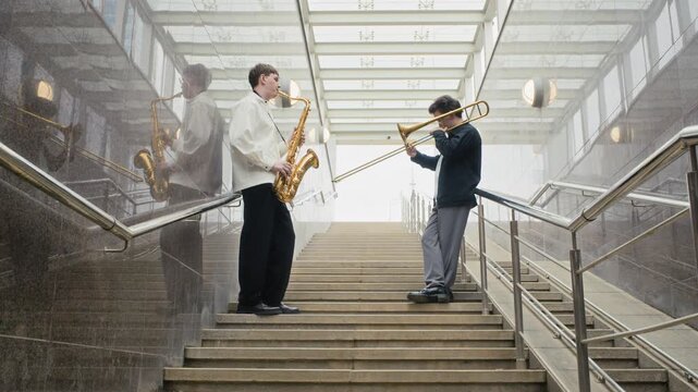 Caucasian young adult musicians playing saxophone and trombone on urban stairs from low angle. Clip fits youth culture campaigns, art documentaries, and music festival advertising