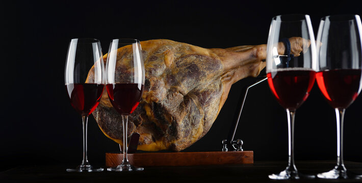 Whole cured ham leg on wooden stand. Four wine glasses filled with red wine. Dark background. Gourmet Spanish delicacy. Wine tasting. Narrow banner