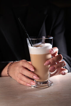 Beautiful female hands are holding a tall glass with a coffee drink. A girl drinks a latte. Latte coffee with foaming milk in a tall glass. Dark background. Hands in close-up