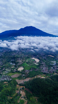 Aerial view of Mount Gede Pangrango National Park rising above a sea of clouds, with lush green landscapes, rice fields, and residential areas in Sukalarang, West Java, Indonesia.