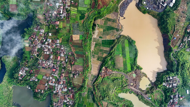 Aerial view of Cimangkok Sand Mining Site with large pits and sediment-filled water basins surrounded by lush green agricultural fields and a village in Sukalarang, West Java, Indonesia.