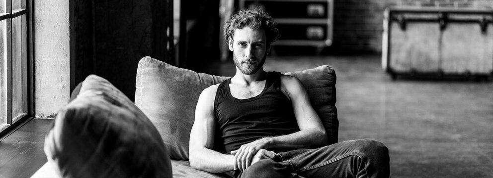Portrait of a fit slim handsome young stylish sexy man in a black t-sjirt shirt, jeans posing in studio with a loft interior. Black and white. Casual clothing, modern style, men's wear. Relaxed, calm