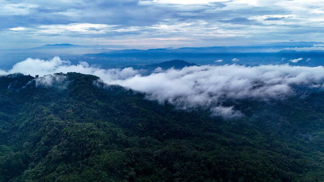 Aerial view of Bukit Sabak Sukabumi featuring lush green forested hills partially covered by low-hanging clouds under a dramatic cloudy sky in Sukalarang, West Java, Indonesia.
