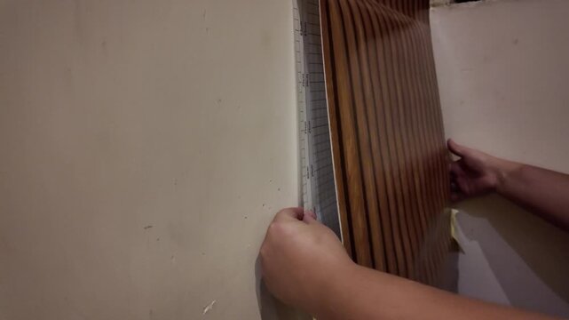 Person installs wood-textured wallpaper on a plain wall. Hands smooth the material with care. Yellow adhesive tape holds edges securely. Ideal for home renovation or interior design projects