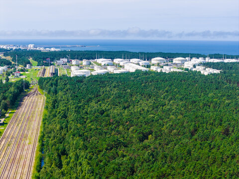Aerial view of the Ventspils oil terminal with numerous storage tanks, dense pine forest, and parallel railway tracks leading towards the Baltic Sea in Ventspils, Latvia.