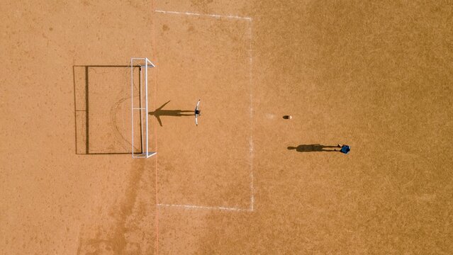 Aerial view of a soccer field with a goalkeeper standing by the goalpost and a player with a ball on the dry, brown ground in Matheran, Maharashtra, India.