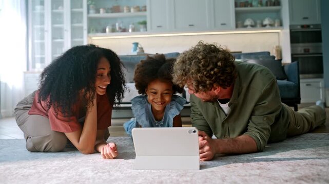 Excited family watching tablet floor kitchen. Mixed race parents laughing home.