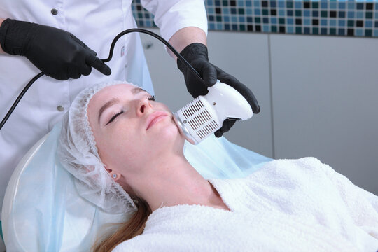 Woman receives facial treatment with white handheld device. Specialist wears black gloves. Clinical skin therapy. Client relaxed on bed. Procedure of hardware facial skin care in cosmetology office