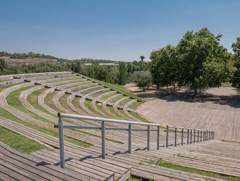 View of the open-air amphitheater in Madrid's Manzanares Linear Park, featuring curved tiered seating with wooden benches and interspersed green spaces, as well as staircases with railings