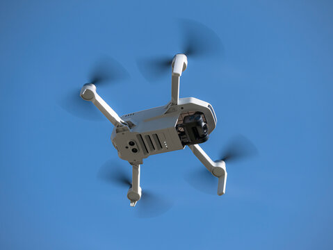 Bottom view of a small white drone with camera flying with its propellers out of focus due to speed flying through the air and a blue sky with clouds in the background