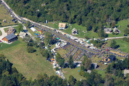 Aerial view of a lively outdoor festival in the Julian Alps with numerous visitors, tents, and dining tables set up in a grassy field near Bohinjsko jezero, Radovljica, Slovenia.