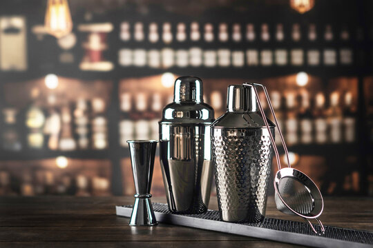 Two metal shakers with jigger and strainer on black rubber mat. Wooden bar top. Blurred bottles background