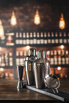 Two cocktail shakers jigger strainer on black mat. Wooden bar counter blurred bottles background
