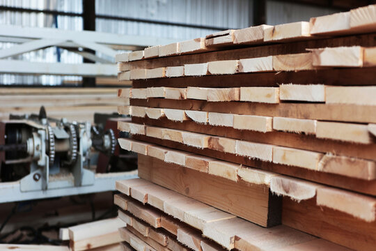 Stack of wooden planks next to an industrial woodworking machine. Grain of wood is visible on ends of planks. Chainsaw is blurred in background. Interior of sawmill