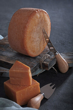 Hard cheese wheel on wooden board with knives, cut wedges. Dark rustic still life. Gourmet dairy product