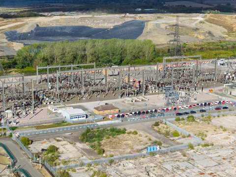 Aerial view of an electrical substation featuring transformers, switchgear, and power lines, with a parking lot containing cars and an industrial landscape in the background, United Kingdom.