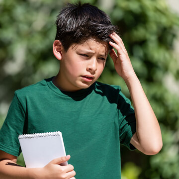 Confused boy in green t-shirt scratches head holding notebook outdoors. Education concept, school, homework, thinking, doubt, problem solving, learning difficulty and puzzled student emotion