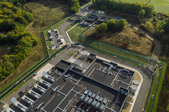 Aerial view of a modern data center facility featuring extensive rooftop cooling infrastructure, a secure perimeter fence, and a paved parking area surrounded by lush woodland in the United Kingdom.
