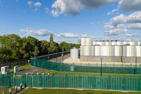 Aerial view of a modern data center facility surrounded by high green security fencing and lush trees under a blue sky with scattered clouds in the United Kingdom.