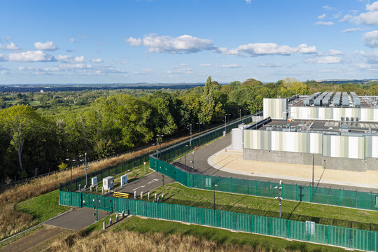 Aerial view of a modern data center facility surrounded by security fencing and lush green trees in the United Kingdom.
