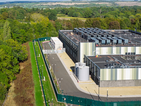 Aerial view of a modern data center facility featuring large cooling units on the roof, storage tanks, and a secure perimeter fence surrounded by lush green trees in the United Kingdom.