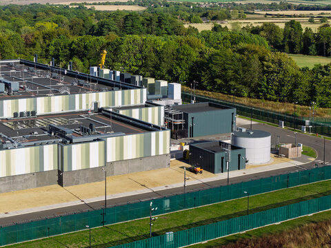 Aerial view of a modern data center facility with industrial rooftops, cooling units, and a storage tank, surrounded by a security fence and lush green trees in the United Kingdom.