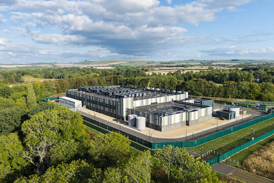Aerial view of a modern data center facility surrounded by lush green trees and open fields under a blue sky in the United Kingdom.