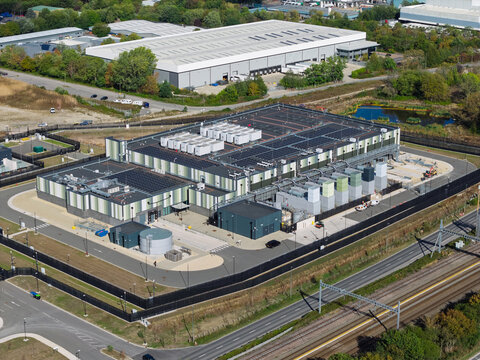 Aerial view of a modern data center facility with rooftop cooling units, surrounded by security fencing, adjacent to a railway line and industrial buildings in the United Kingdom.