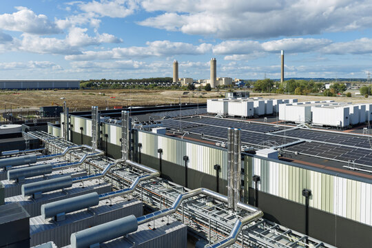 Aerial view of a modern data center facility featuring extensive rooftop solar panels, complex cooling infrastructure with metallic pipes, and industrial chimneys in the background, United Kingdom.