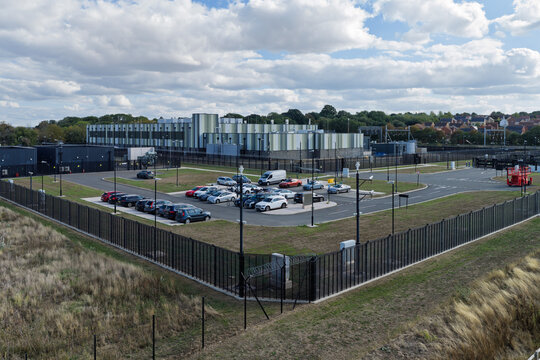 Aerial view of a modern data center facility with a parking lot, security fencing, and surrounding green landscape in the United Kingdom.