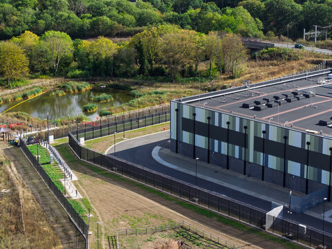 Aerial view of a modern data center facility with a fenced perimeter, a small pond, and surrounding greenery in the United Kingdom.