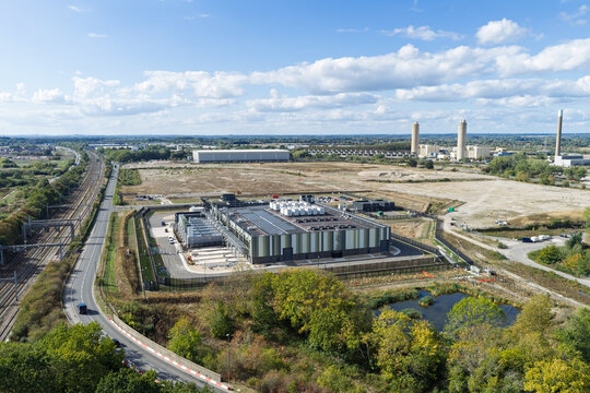 Aerial view of a modern data center facility surrounded by open fields, a railway line, and a road in the United Kingdom.