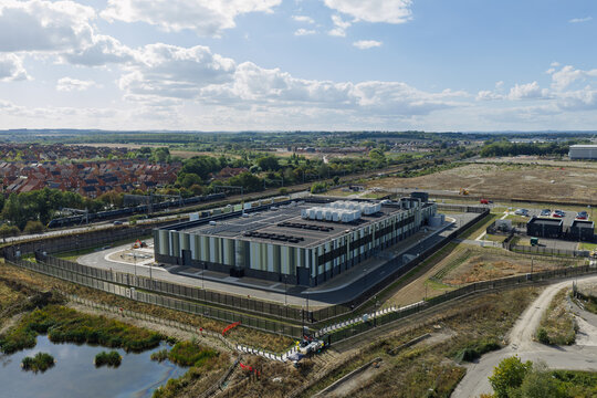 Aerial view of a modern data center facility with rooftop cooling units, surrounded by security fencing, adjacent to a railway line and a pond in the United Kingdom.