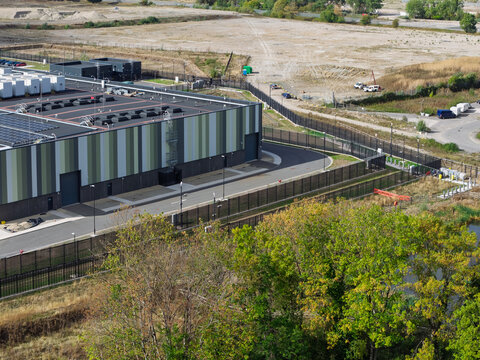 Aerial view of a modern data center building with solar panels on the roof, surrounded by a security fence and green trees in the United Kingdom.
