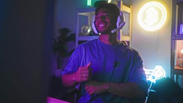 Man wearing headset claps hands and smiles broadly in studio with neon bitcoin sign, shelving and a game controller visible on the desk; joyful energy.