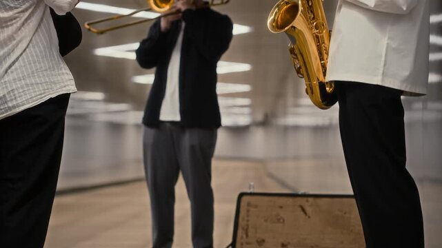 Caucasian young adult musicians playing saxophone and trombone in pedestrian tunnel with passerby dropping money into open case. Clip ideal for urban lifestyle and music industry storytelling