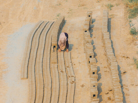 Aerial view of a worker arranging rows of sun-dried mud bricks in a dusty open-air factory in Lahore, Punjab, Pakistan.