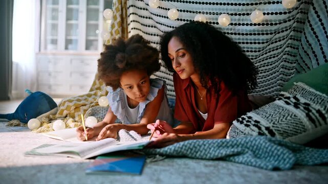 Mom helping schoolgirl homework blanket fort. Family drawing together indoors.